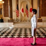 A woman in formal attire standing in a grand hall with flags in the background