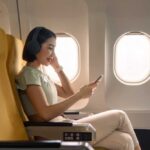 A woman sitting in an airplane seat, wearing headphones and using her phone