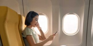 A woman sitting in an airplane seat, wearing headphones and using her phone
