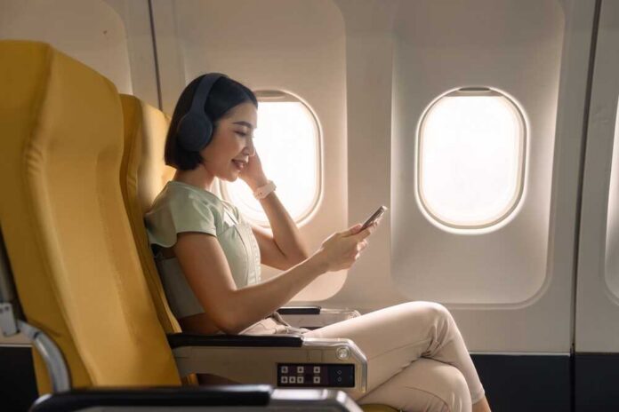 A woman sitting in an airplane seat, wearing headphones and using her phone