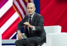 A man in a suit speaking at a conference with an American flag in the background