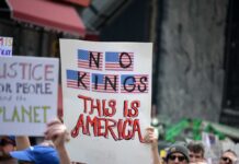 A protester holding a sign that reads 'NO KINGS THIS IS AMERICA' with American flags