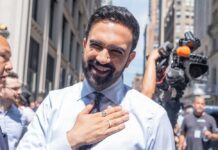 A smiling man in a light blue shirt gestures with his hand in an outdoor urban setting