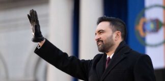 A man in a black coat and gloves waving during a formal event