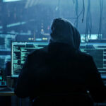 A person in a hoodie working on multiple computer screens in a dimly lit room