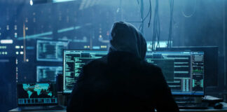 A person in a hoodie working on multiple computer screens in a dimly lit room