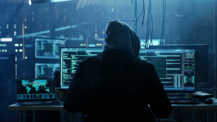 A person in a hoodie working on multiple computer screens in a dimly lit room