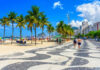 A coastal walkway lined with palm trees and beachgoers enjoying a sunny day