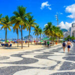 A coastal walkway lined with palm trees and beachgoers enjoying a sunny day