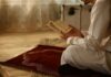 Person in white attire reading a religious text while kneeling on a prayer rug