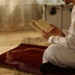 Person in white attire reading a religious text while kneeling on a prayer rug