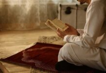 Person in white attire reading a religious text while kneeling on a prayer rug