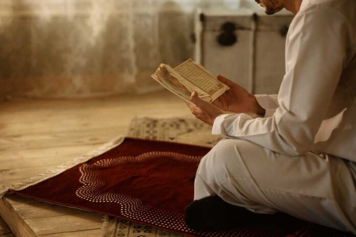 Person in white attire reading a religious text while kneeling on a prayer rug