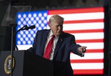 A man in a suit gestures while speaking at a podium with an American flag in the background
