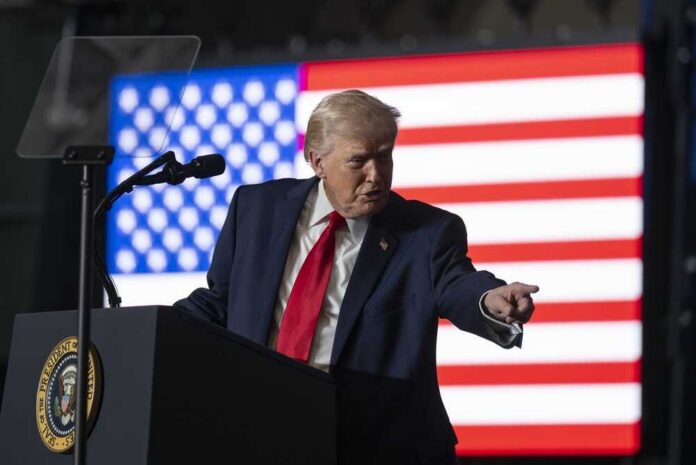 A man in a suit gestures while speaking at a podium with an American flag in the background