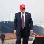 A man in a suit wearing a red cap stands near a ship at a harbor