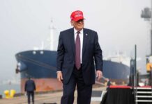 A man in a suit wearing a red cap stands near a ship at a harbor