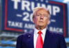 man in suit with red tie standing before a blurred campaign-style sign