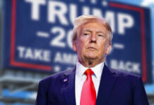 man in suit with red tie standing before a blurred campaign-style sign