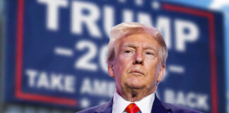 man in suit with red tie standing before a blurred campaign-style sign