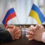 Two pairs of hands clasped together with Russian and Ukrainian flags in the background