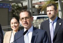 A man in a suit speaking at a press conference with two others in the background