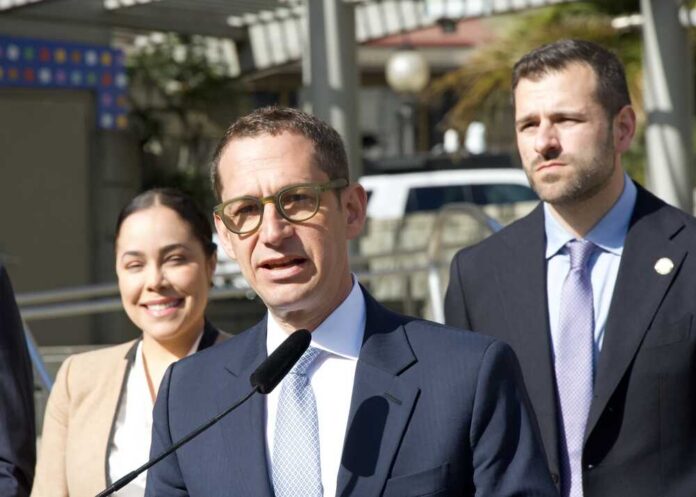 A man in a suit speaking at a press conference with two others in the background