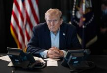 A serious-looking man seated at a table with phones and an American flag in the background