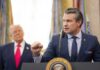 A man in a suit delivering a speech at a podium with a presidential seal, while another man stands in the background.