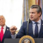 A man in a suit delivering a speech at a podium with a presidential seal, while another man stands in the background.