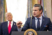 A man in a suit delivering a speech at a podium with a presidential seal, while another man stands in the background.