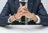 A man in a suit with hands handcuffed together on a table