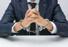 A man in a suit with hands handcuffed together on a table