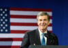 A smiling politician speaking at a podium with an American flag in the background