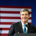 A smiling politician speaking at a podium with an American flag in the background