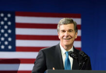 A smiling politician speaking at a podium with an American flag in the background