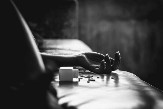 Hand on bed beside spilled pill bottle and tablets