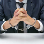 A man in a suit with hands handcuffed together on a table