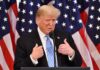 man in suit speaking at podium with US flags behind