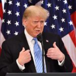 man in suit speaking at podium with US flags behind