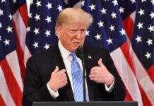 man in suit speaking at podium with US flags behind