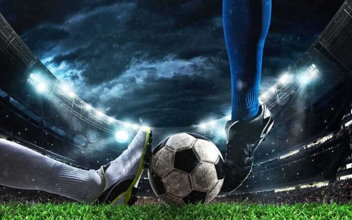 shutterstock_1937437555 soccer players' feet contest the ball in a lit stadium at night