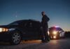 Police officer stands beside a stopped car at dusk