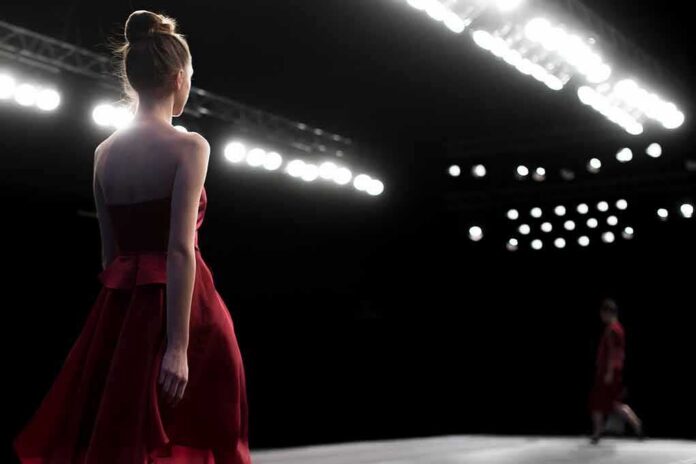 Model in a red dress walking on a runway under bright stage lights
