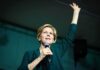 Elizabeth Warren speaking on stage with a microphone, raising one hand
