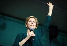 Elizabeth Warren speaking on stage with a microphone, raising one hand
