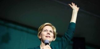 Elizabeth Warren speaking on stage with a microphone, raising one hand
